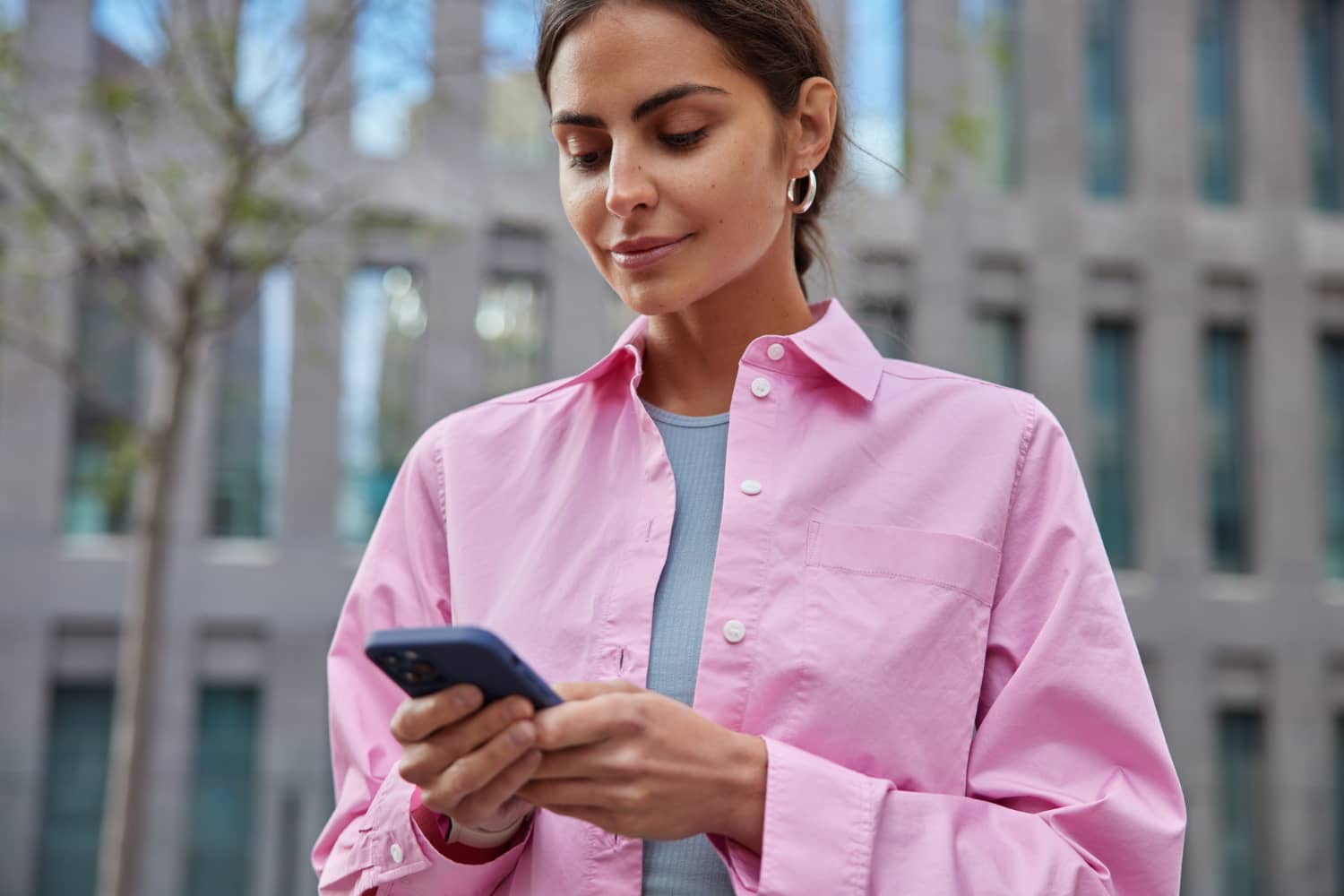 horizontal shot beautiful millennial girl uses mobile phone city finding route explores new places interest wears pink shirt poses blurred building